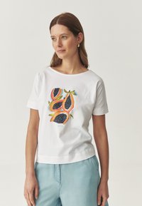 Hvid bomulds t-shirt med et farverigt frugtdesign i orange og blå toner. Korte ærmer og en afslappet pasform. Lyseblå shorts synlige.