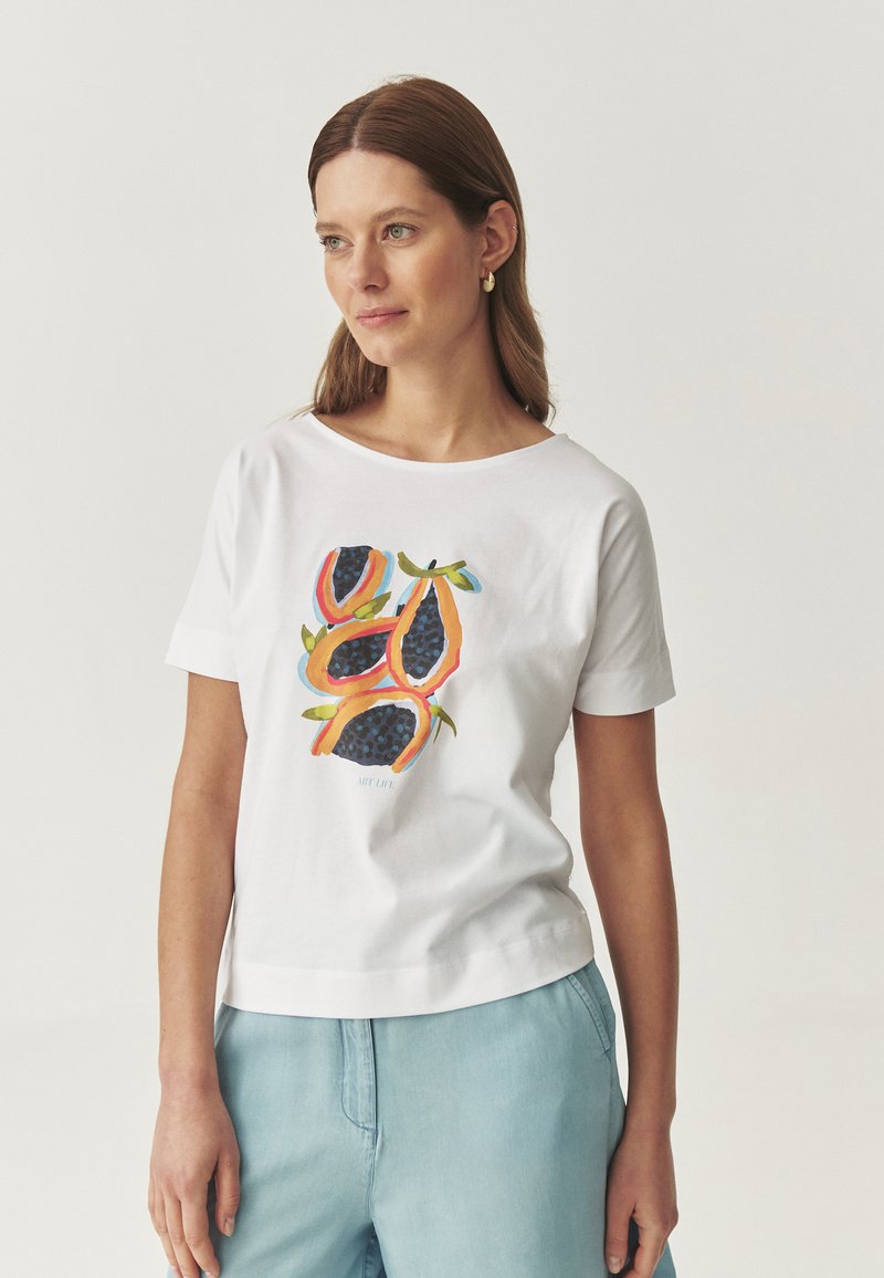 Hvid bomulds t-shirt med et farverigt frugtdesign i orange og blå toner. Korte ærmer og en afslappet pasform. Lyseblå shorts synlige.