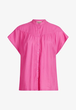 Kobieta w jasnoróżowej bluzce na guziki z krótkim rękawem, włożonej do jasnoniebieskich dżinsów z efektem sprania w stylu acid wash z wysokim stanem, z rękami w kieszeniach.