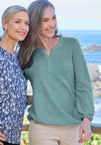 Due donne stanno insieme all'aperto. Una indossa un pullover teal con scollatura henley; l'altra ha una maglietta ampia a fantasia nei toni del blu e del viola.