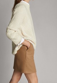 Pull en maille crème avec un motif texturé, associé à une jupe moulante marron clair dotée de poches avant. Col de chemise blanc visible.