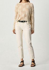 Pull en maille beige à motif ajouré, jeans crème et ceinture noire. Porté avec des sandales noires à brides, avec semelles plates et brides à la cheville.