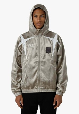 Giovane uomo che indossa una giacca con cappuccio color argento lucido con logo e stemma Umbro bianchi, chiusa con la cerniera, in piedi davanti a uno sfondo semplice.