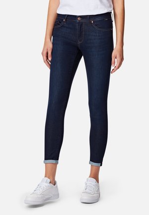 Person wearing dark blue rolled-up skinny jeans and white sneakers, standing with hands relaxed by sides.