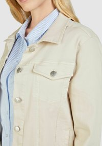 Beige Jeansjacke mit silbernen Druckknöpfen, einer Brusttasche und einem spitzen Kragen. Getragen über einem hellblauen und weißen gestreiften Hemd.