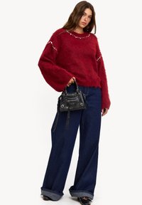 Pull en peluche rouge avec des manches oversized, orné d'accents argentés. Assorti à un jean évasé bleu foncé et à un petit sac à main noir.