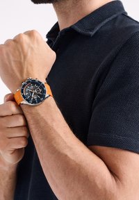 Uomo che indossa una polo in tessuto blu navy, mostrando un orologio con quadrante nero e cinturino in gomma arancione alzando il braccio.