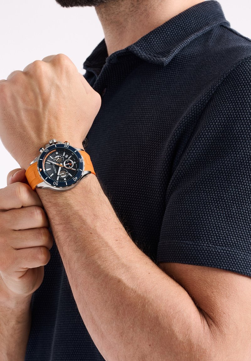 Uomo che indossa una polo in tessuto blu navy, mostrando un orologio con quadrante nero e cinturino in gomma arancione alzando il braccio.