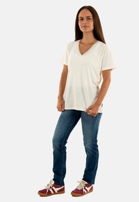 Camiseta blanca de cuello en V con mangas cortas, combinada con jeans azules y zapatillas burdeos con detalles blancos, posando frente a un fondo liso.