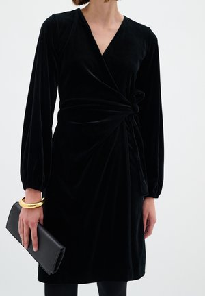 Black velvet wrap dress with long puff sleeves, featuring a waist tie and a deep V-neck. Accessories include a black clutch and gold bracelet.