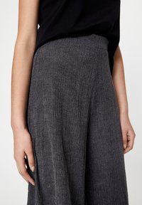 Szerokie, teksturowane spodnie w ciemnoszarym materiale, z wysokim stanem i subtelnym wykończeniem z krepy. Ręka spoczywająca obok spodni.