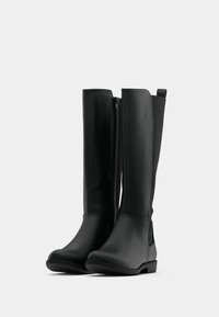 Čierne čižmy po kolená z hladkého syntetického materiálu, so zipsom na boku, plochým podpätkom a minimalistickým dizajnom. Okrúhla špička a jemné detaily.