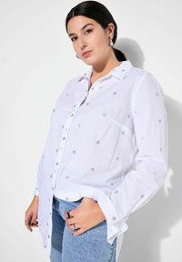 Weißes Hemd mit Knöpfen, das silberne Herzmuster aufweist, lange Ärmel hat und locker sitzt, kombiniert mit hellblauen Jeans.