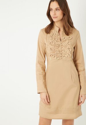 Vestido beige de manga larga, con un patrón floral texturizado y cierre de botones, acentuado por costuras decorativas a lo largo del dobladillo.