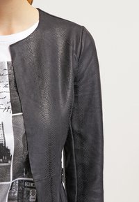 Donna che indossa una giacca in pelle testurizzata nera sopra una t-shirt bianca con grafiche che ritraggono immagini della città in bianco e nero e scritte.