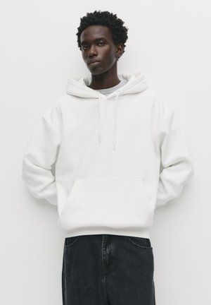 Hvid hættetrøje med frontlomme, snore og oversized pasform. Kombineret med mørk denim, der fremviser en blød, glat stofstruktur.