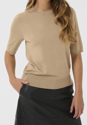 Femme portant un haut en maille beige à manches courtes et une jupe en cuir noire, une main dans la poche de la jupe, sur un fond uni.