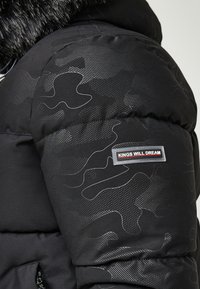 Chaqueta acolchada negra con un patrón de camuflaje y acentos texturizados. Presenta un parche de marca con "KINGS WILL DREAM" en rojo y gris.