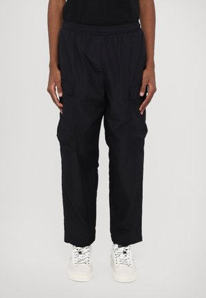 Personne portant un pantalon cargo noir avec des poches à rabat à l'avant et des baskets blanches, debout devant un fond uni.