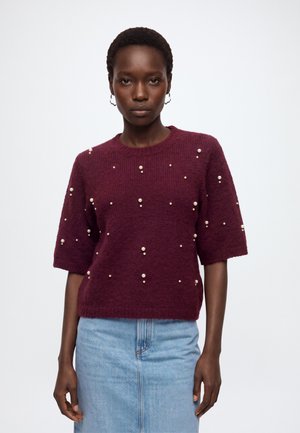 Femme portant un pull bordeaux à manches courtes avec de petites décorations blanches, associé à une jupe en denim bleu clair, debout devant un fond blanc.