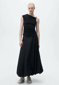 Top negru fără mâneci cu design plisat și accesorii din metal argintiu, asortat cu o fustă lungă neagră. Material neted, siluetă lejeră, încălțăminte casual.