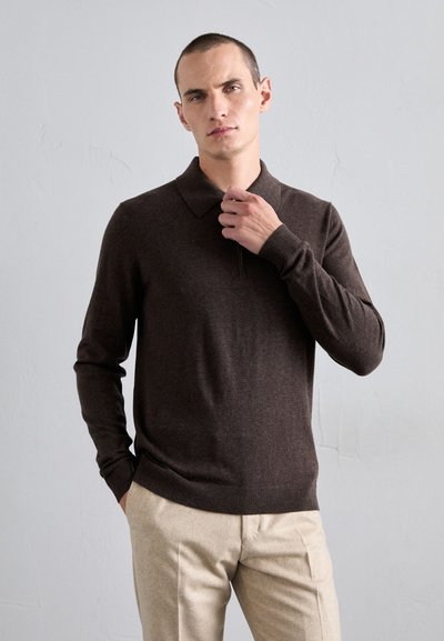 Hombre vistiendo un suéter de polo de manga larga marrón oscuro y pantalones beige, con una mano en el bolsillo y la otra ajustándose el collar, fondo neutral.