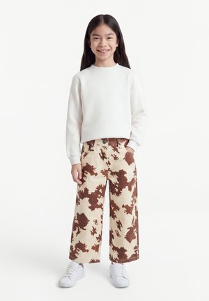 Fille souriante aux longs cheveux foncés, portant un sweat blanc, un pantalon large marron et beige à motifs et des baskets blanches, debout les mains dans les poches.
