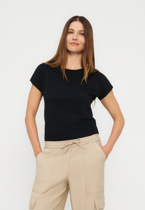 Femme aux cheveux longs, raides et bruns, portant un haut noir à manches courtes et un pantalon beige à cordon, debout les mains dans les poches.