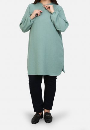 Woman wearing a loose, long-sleeve sage green tunic, black pants, and black loafers, adjusting a necklace against a plain light background.