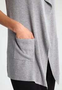 Šedý cardigan s jemnou texturou, s hlubokým véčkovým výstřihem, předními kapsami a asymetrickým lemem. Nositý přes černé kalhoty.