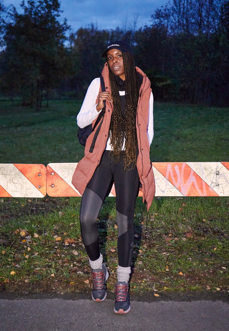 Brązowa bezrękawnik puchowy na białej koszuli z długim rękawem, czarne leginsy i szare skarpetki. Ciemne sneakersy. Stoi przy pasie z paskami.