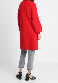 Rode gebreide lange trui met ribbeltextuur, oversized mouwen en zijsplitten, gecombineerd met grijze jeans en zwarte chunky hakken sandalen.