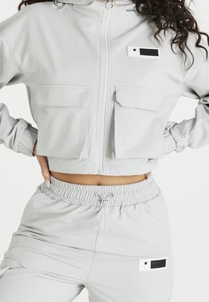 Vrouw draagt een lichtgrijze cropped jas met ritssluiting en grote voorzakken en bijpassende broek met trekkoord en elastische taille.