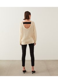 Suéter de punto crema con diseño de espalda descubierta, llevado sobre una blusa negra, combinado con pantalones ajustados negros y tacones negros. Textura suave, corte relajado.