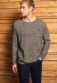 Muž v šedém texturovaném svetru s dlouhým rukávem a tmavě modrých džínách stojí proti horizontální dřevěné ploše a dívá se vlevo.