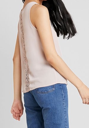 Vrouw draagt een mouwloze beige top met kantdetail aan de achterkant, gecombineerd met blauwe spijkerbroek, staand tegen een effen achtergrond.