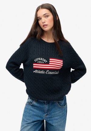 Marineblauwe kabelgebreide trui met een ronde hals, voorzien van een rood-wit gestreepte graphic en de tekst "Athletic Essentials" op de voorkant.