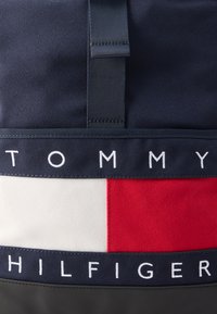 Σκούρο μπλε ύφασμα σακίδιο πλάτης με ο ριζόντια λουράκι που φέρει λευκά γράμματα "TOMMY" και "HILFIGER" και κόκκινο-λευκή διακοσμητική λεπτομέρεια.