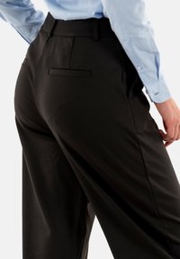 Pantaloni neri a vita alta con una texture liscia, dotati di una tasca posteriore e dettagli di cucitura discreti. Abbinati a una camicia azzurro chiaro.