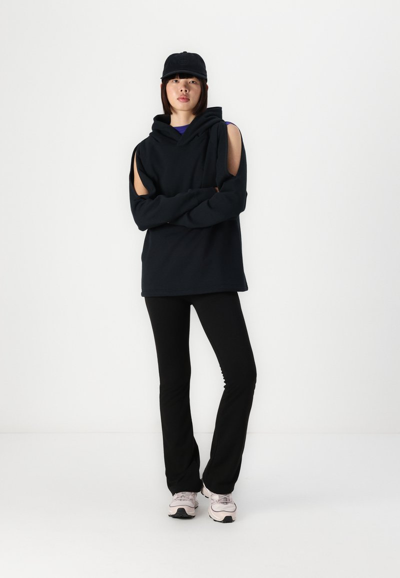 Felpa nera con spalle scoperte, indossata sopra un top viola, abbinata a pantaloni neri e sneaker grigie. Gli accessori comprendono un cappellino nero.