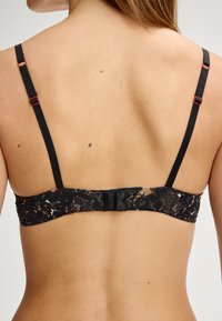 Czarny koronkowy biustonosz z regulowanymi ramiączkami, z teksturowanym wzorem w odcieniach czerni i beżu. Zawiera zapięcie na haczyk z tyłu.