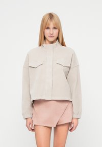 Veste en velours côtelé beige raccourcie avec col montant, deux poches poitrine et poignets élastiqués, associée à une mini-jupe en suède rose clair.