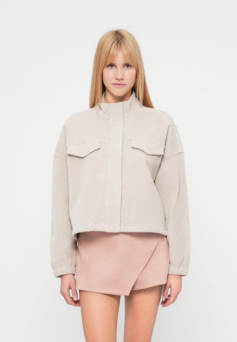 Veste en velours côtelé beige raccourcie avec col montant, deux poches poitrine et poignets élastiqués, associée à une mini-jupe en suède rose clair.