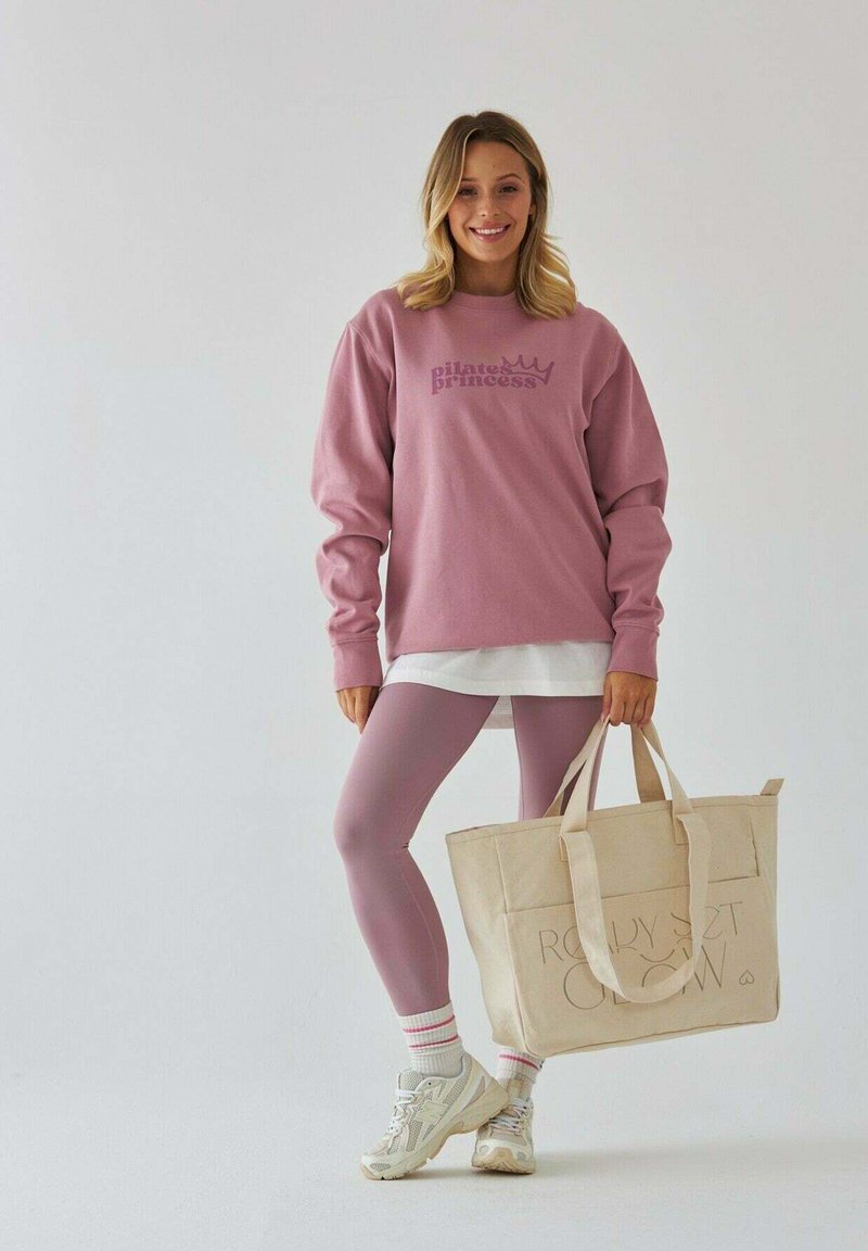 Różowy sweatshirt z napisem "Pilates Princess", w połączeniu z dopasowanymi legginsami, białą warstwową bluzką oraz beżową torbą. Całość dopełniają białe sneakersy.