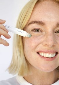 Sérum pour les yeux à base d'eau de raisin dans un tube transparent avec un applicateur roll-on. L'utilisateur l'applique sur la zone sous les yeux. Peau et cheveux clairs.