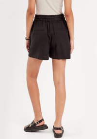 Shorts en lin noir avec une taille à cordon, deux poches arrière et une coupe décontractée. Portés avec des sandales noires à lanières.