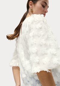 Witte textuurblouse met bloemenapplicaties, pofmouwen en een ronde halslijn, gecombineerd met gouden hoepelringen.