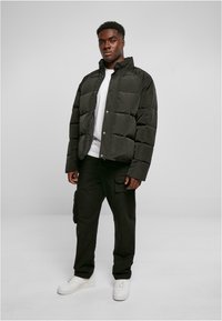 Chaqueta tipo plumas en verde oscuro con patrón acolchado, cierre de botones a presión y cuello alto. Llevada con pantalones cargo negros y deportivas blancas.