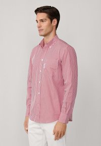 Homme portant une chemise à rayures verticales rouges et blanches à manches longues avec poche avant, associé à un pantalon blanc, se tenant devant un fond uni.