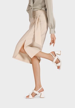 Gonna midi beige con dettagli in vita legati e spacco laterale, abbinata a sandali con tacco blocco bianchi caratterizzati da accenti rotondi e cinturini alla caviglia.
